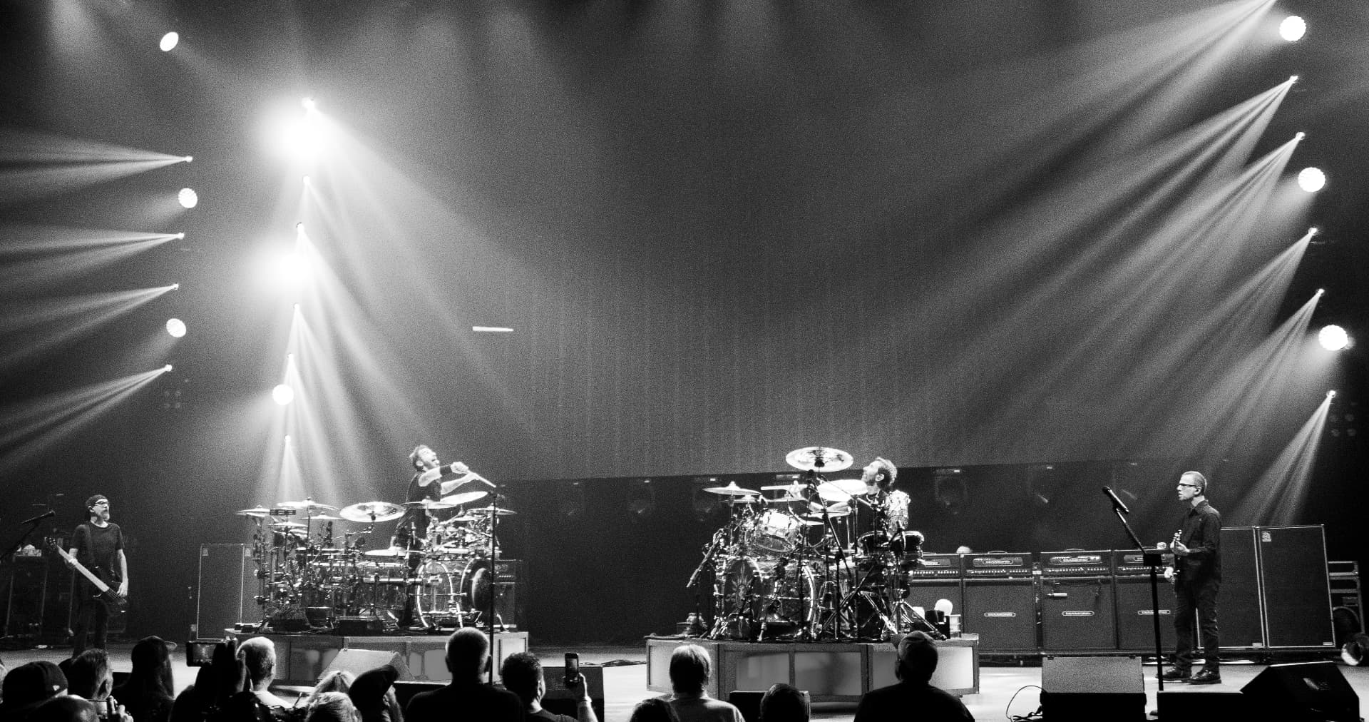 Wide black and white arena stage with two drum kits and beams of light